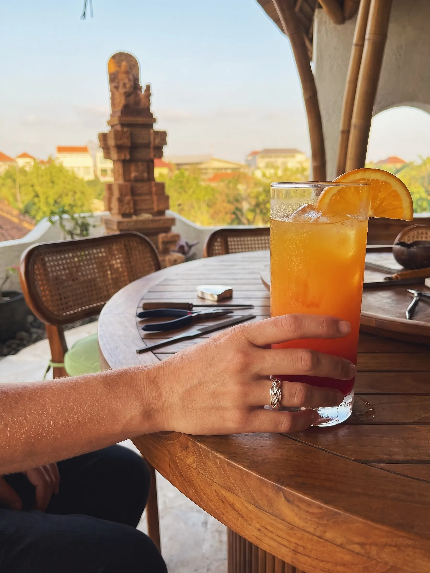A guest wearing a braided silver ring holds a Bali Sunset drink at the teak bench of the open-air gallery